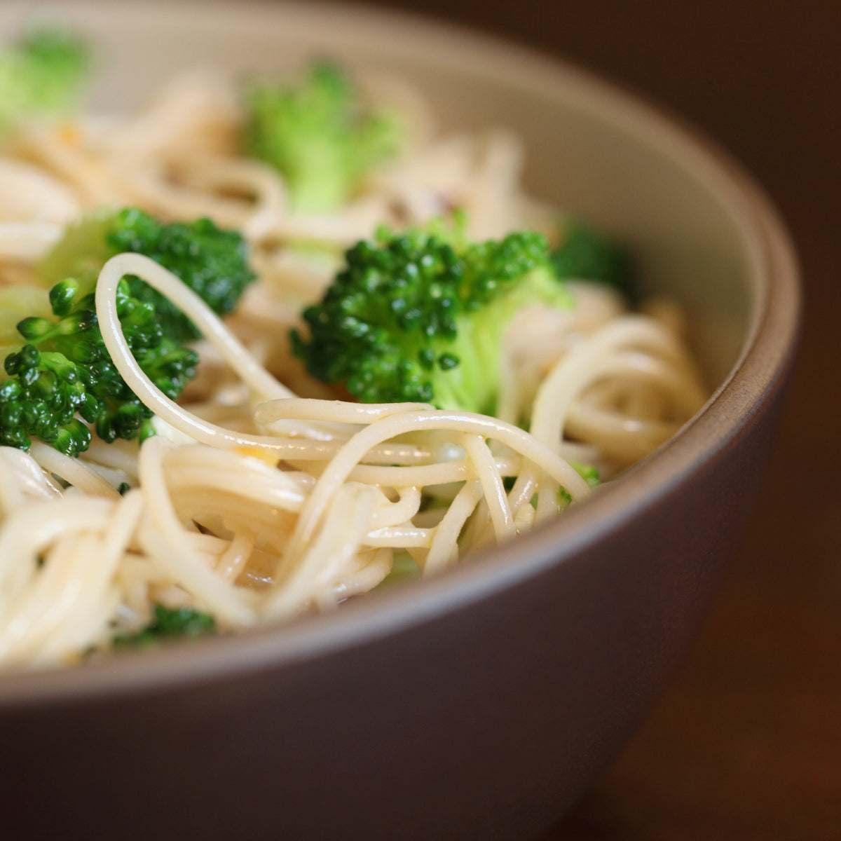 Bowl of cooked brown rice capellini tossed with broccoli florets, served in a brown bowl on a wooden surface