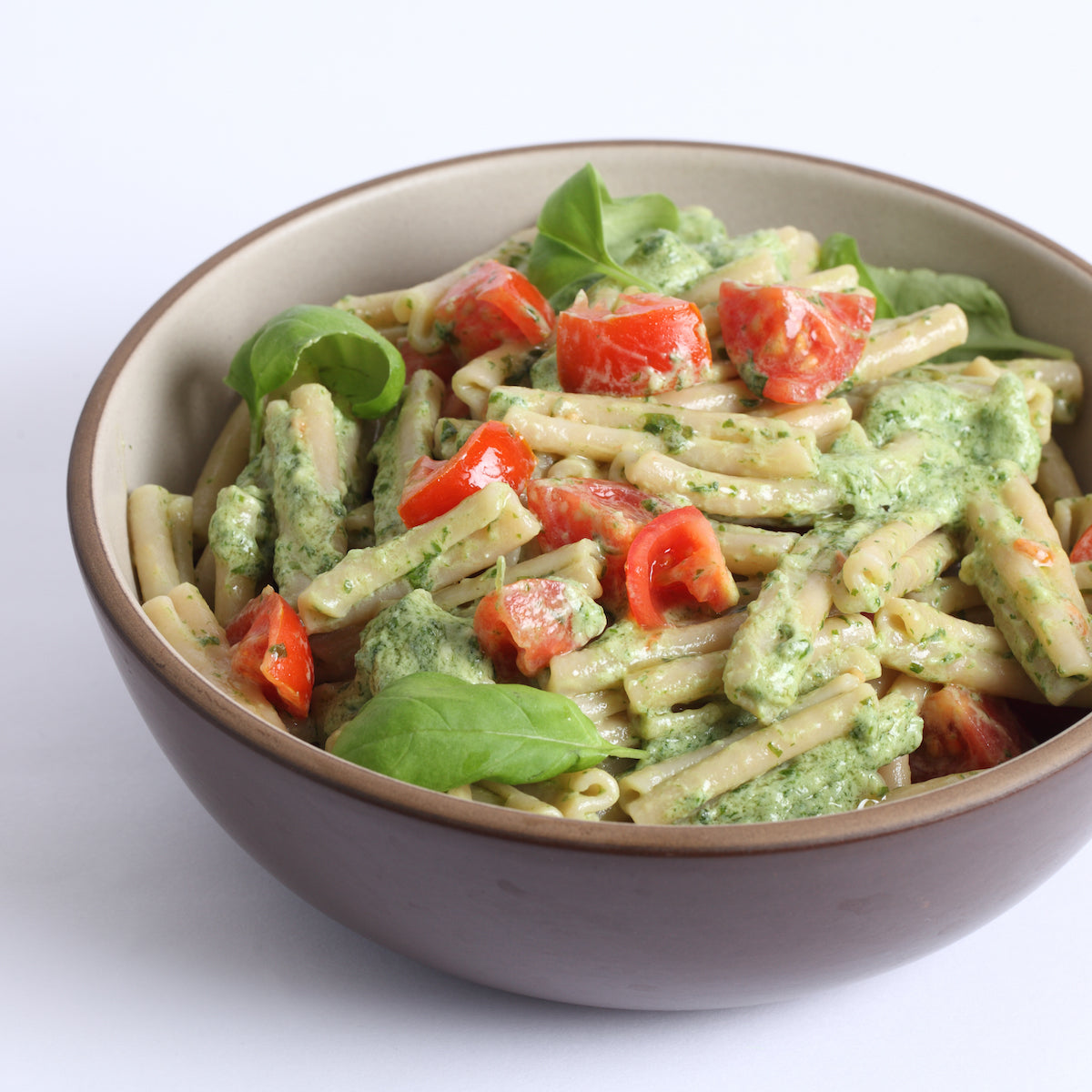 Bowl of brown rice caserecce tossed in pesto with cherry tomatoes and basil on a white background