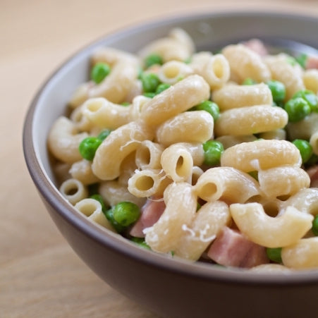 Close-up of cooked Brown Rice Elbows showcasing firm texture with peas and diced ham in a shallow beige bowl on a wooden surface