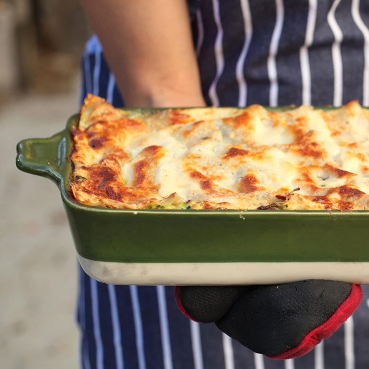Baked organic gluten-free brown rice lasagna in a green ceramic baking dish held by someone in a striped apron and oven mitt