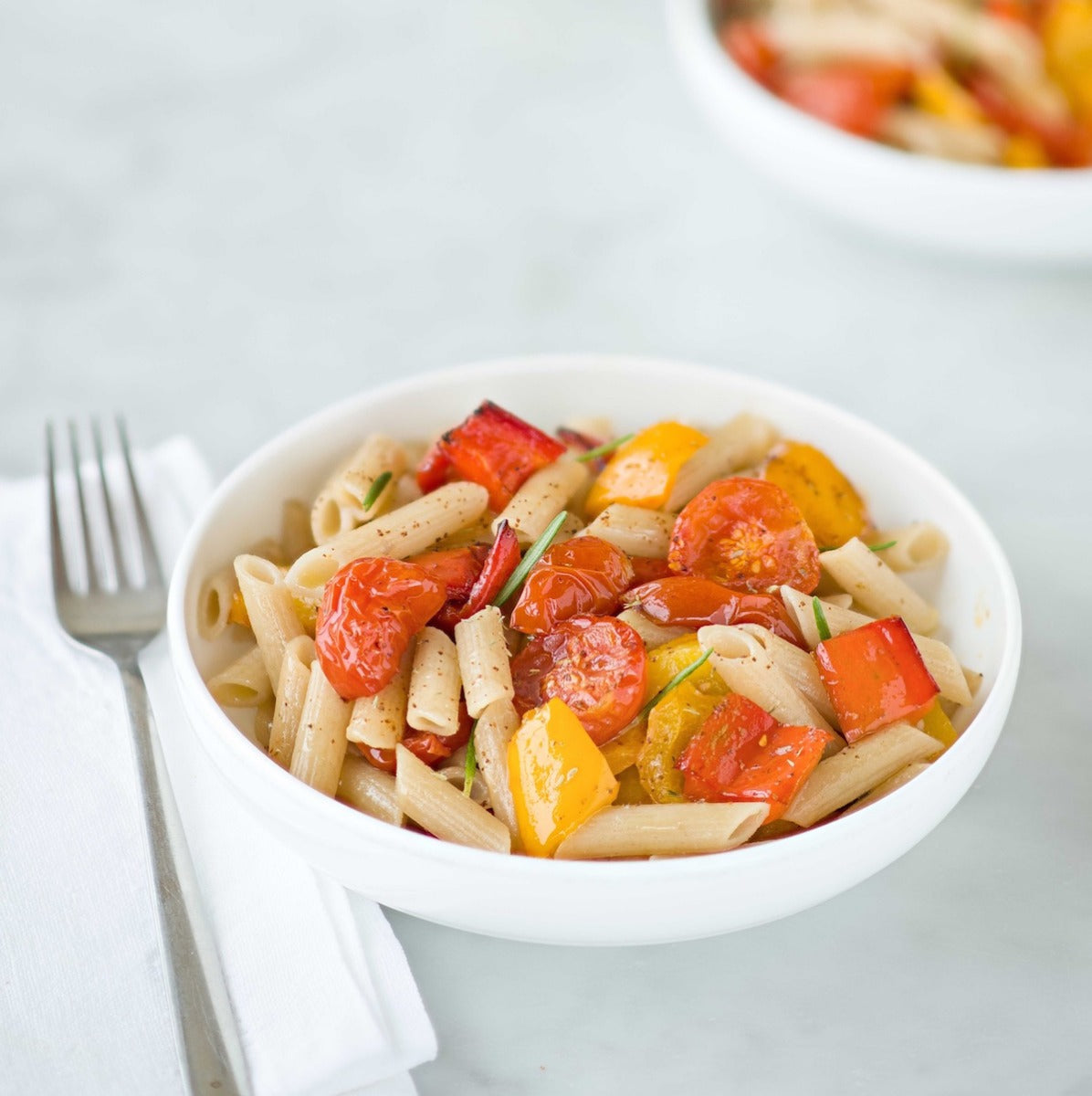 Bowl of organic gluten-free brown rice penne rigate tossed with roasted red and yellow peppers and cherry tomatoes, fork at the side on a light tabletop