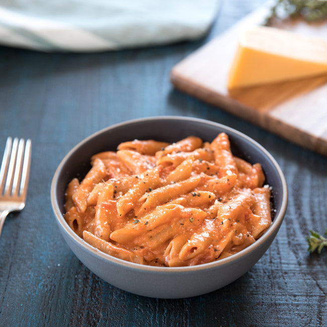 Bowl of Organic Gluten Free Brown Rice Penne in creamy tomato sauce on a dark wood table with a fork and wedge of cheese in the background