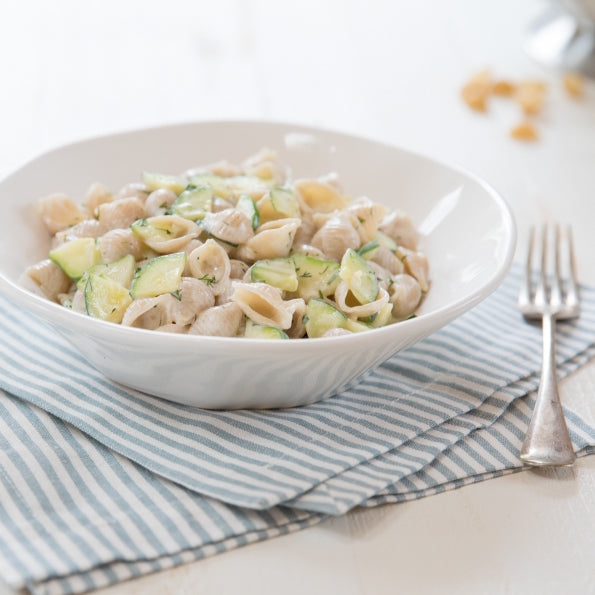 Bowl of Organic Gluten Free brown rice shell pasta tossed with zucchini ribbons and light cream sauce on a blue-striped napkin