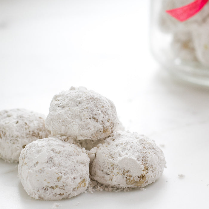 Close-up of four Einkorn walnut snowball cookies with powdered sugar on a white surface and a blurred jar in the background
