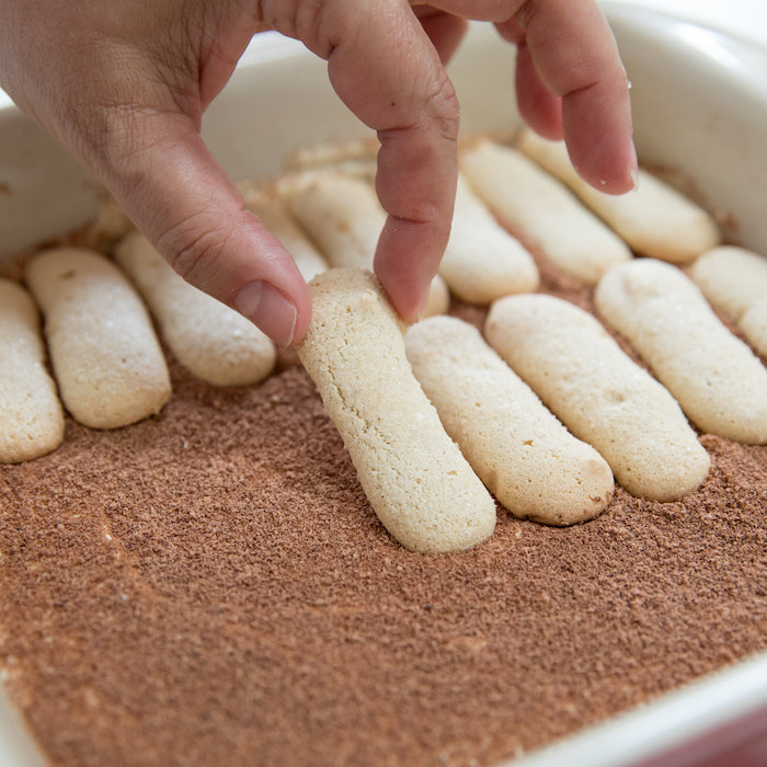 Hand placing Einkorn ladyfinger onto cocoa-dusted tiramisu in a white baking dish