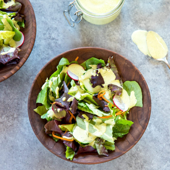 Green Goddess salad dressing drizzled over mixed salad greens with radish and carrot in a wooden bowl, jar and spoon of dressing nearby