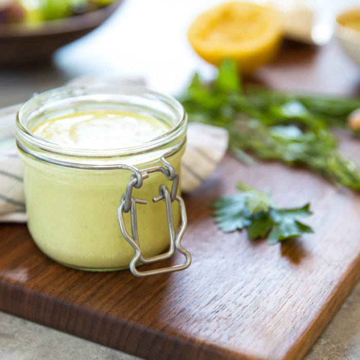 Glass jar of Green Goddess dressing showing creamy light-green texture on wooden board with parsley and lemon