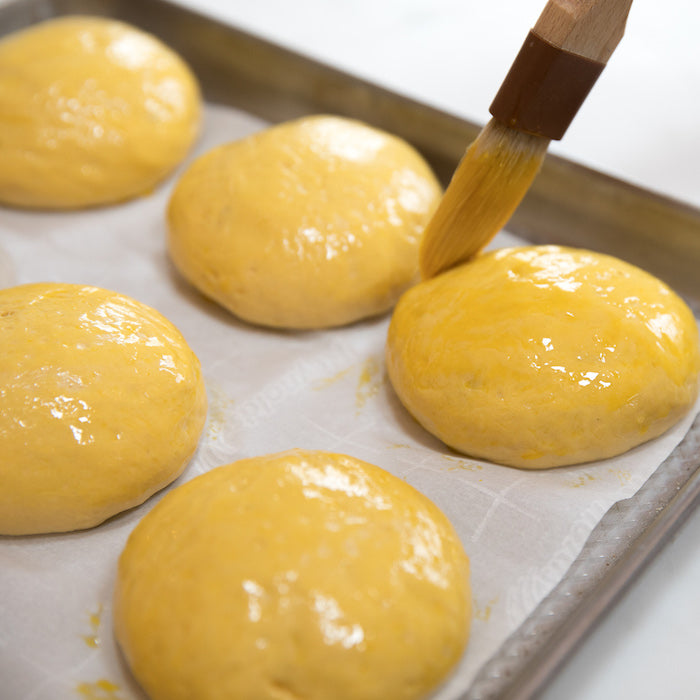 Close-up of raw einkorn hamburger buns on a parchment-lined baking sheet being brushed with egg wash