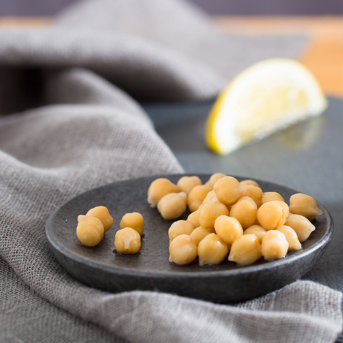 Close-up of chickpeas on a small dark plate showing plump texture with gray linen and a lemon wedge blurred in the background