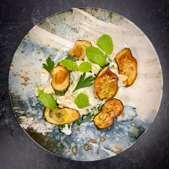 Top-down view of penne with ricotta, lemon, herbs and fried zucchini on a blue-speckled ceramic plate