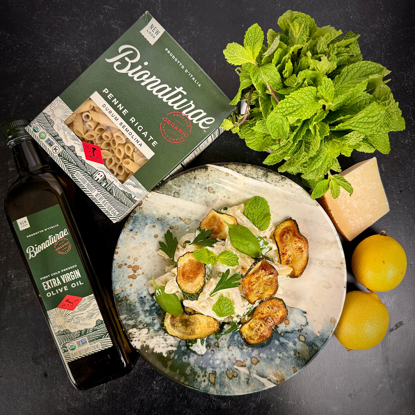 Plated organic penne rigate with herbs served beside a Bionaturae pasta box, olive oil bottle, fresh basil and lemons on a dark tabletop