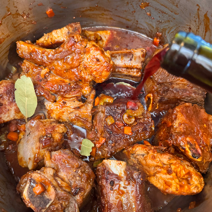 Pork and short rib ragù simmering in a pot with bay leaves and diced carrots and celery as red wine is poured