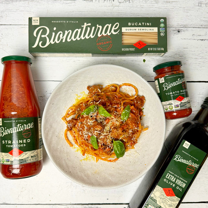 Sausage ragu on bucatini, garnished with basil and cheese, with Bionaturae pasta, tomato jars, and olive oil on white wood