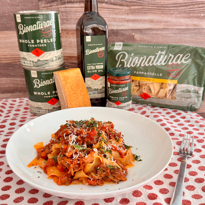 Pappardelle with pork and short rib ragu on a white plate, with Bionaturae packaging, olive oil, canned tomatoes, and cheese