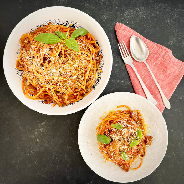 Two bowls of sausage ragu pasta served with grated cheese and basil on a dark surface with a red napkin and utensils