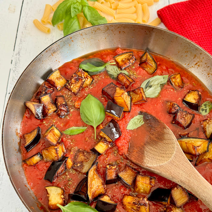 Skillet of Italian tomato sauce with roasted eggplant and fresh basil, wooden spoon in pan, rigatoni and red cloth at top