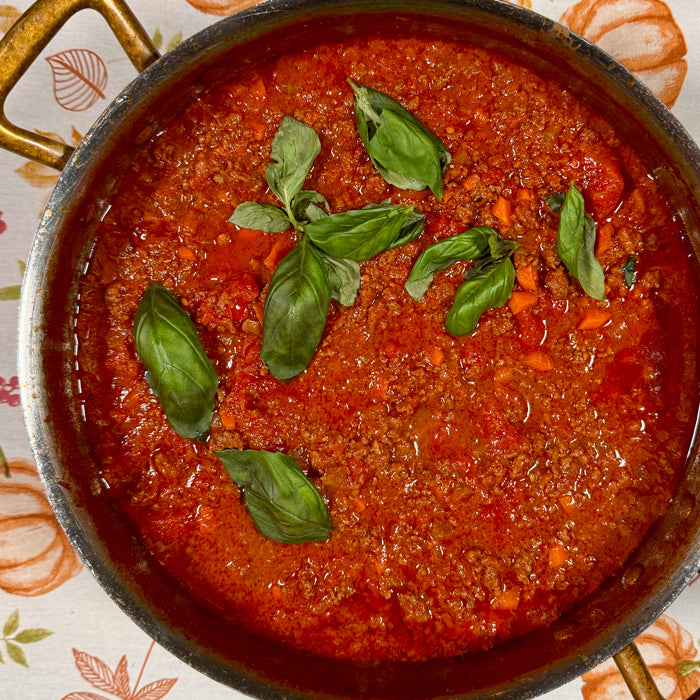 Ragù alla Bolognese simmering in a pot on a patterned tablecloth, topped with fresh basil leaves