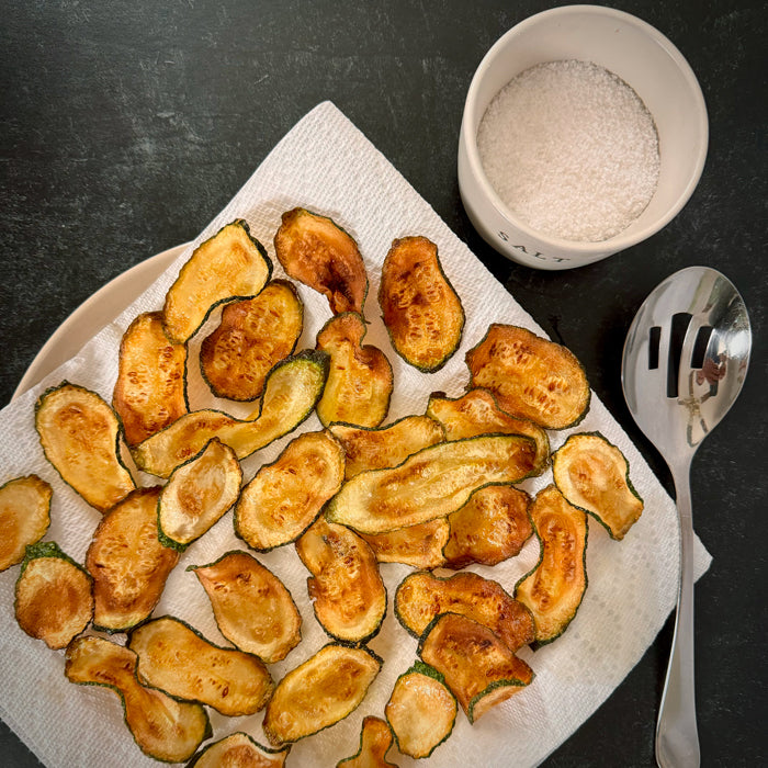 Fried zucchini slices draining on paper towel with a bowl of salt and a slotted spoon on a dark countertop