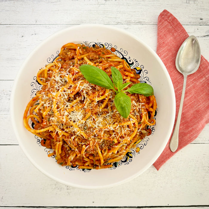 Bowl of spaghetti with pork sausage ragù and grated cheese, garnished with basil on a white wooden table with a red napkin and spoon