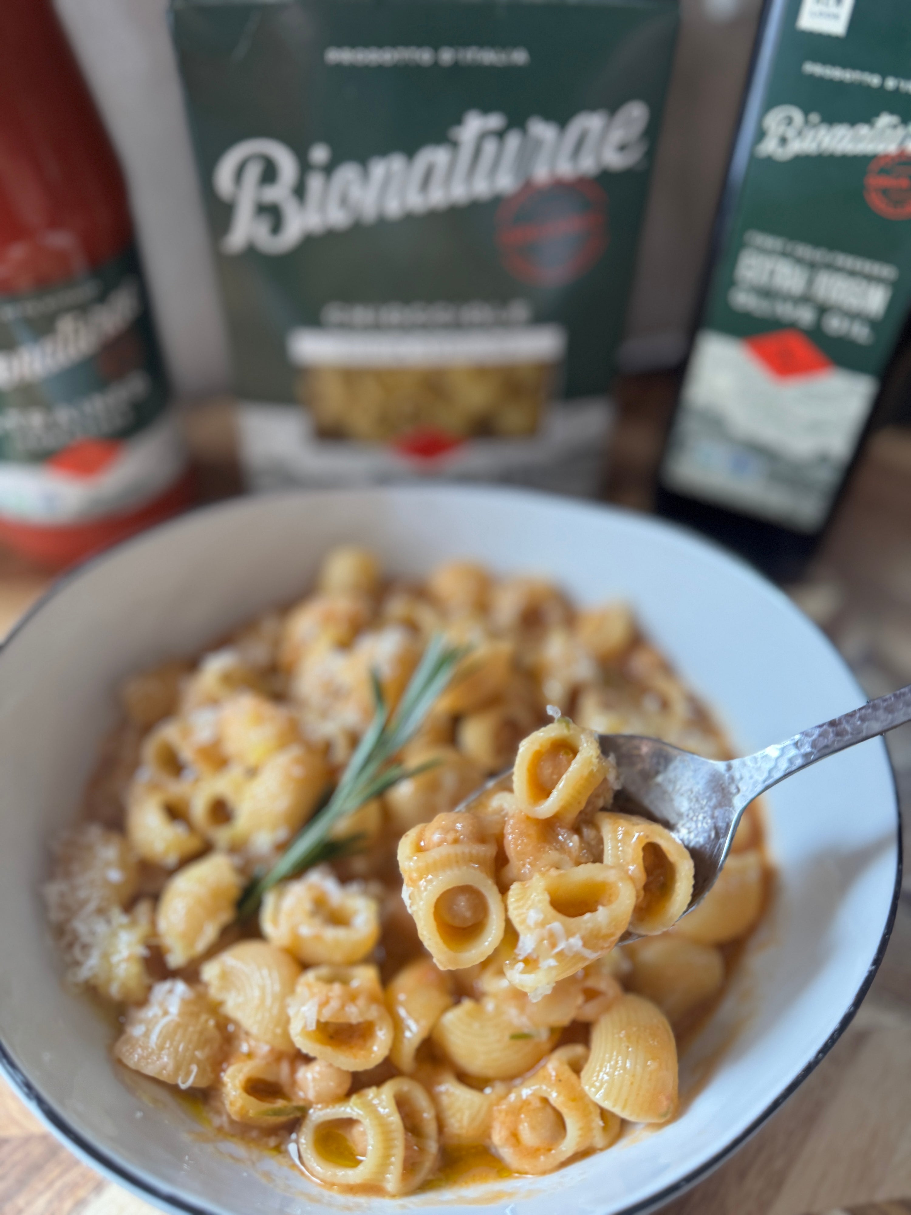 Bowl of chiocciole pasta with chickpeas in tomato broth, fork lifting bite, rosemary garnish, blurred products in background