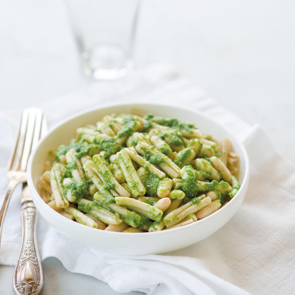 Organic gluten free brown rice caserecce tossed with arugula pesto and white beans in a white bowl on a cloth napkin