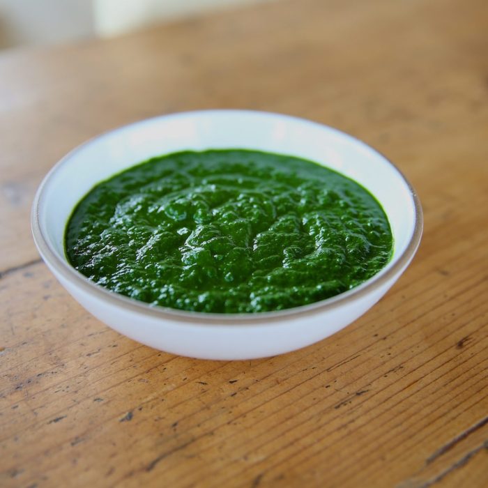 Angled view of a white bowl of organic kale pesto showing vibrant smooth bright green texture on a wooden table