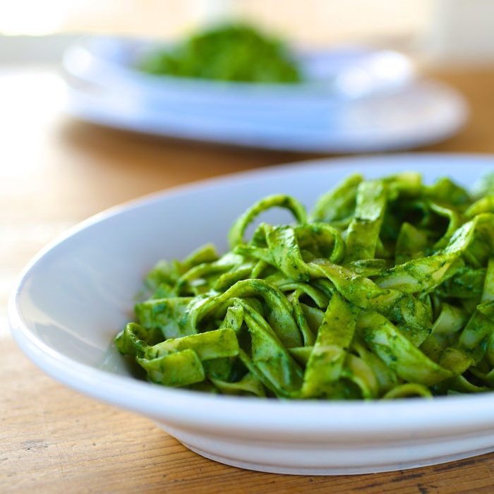 Tagliatelle-style pasta tossed in bright organic kale pesto in a white bowl on a wooden table, second bowl blurred behind