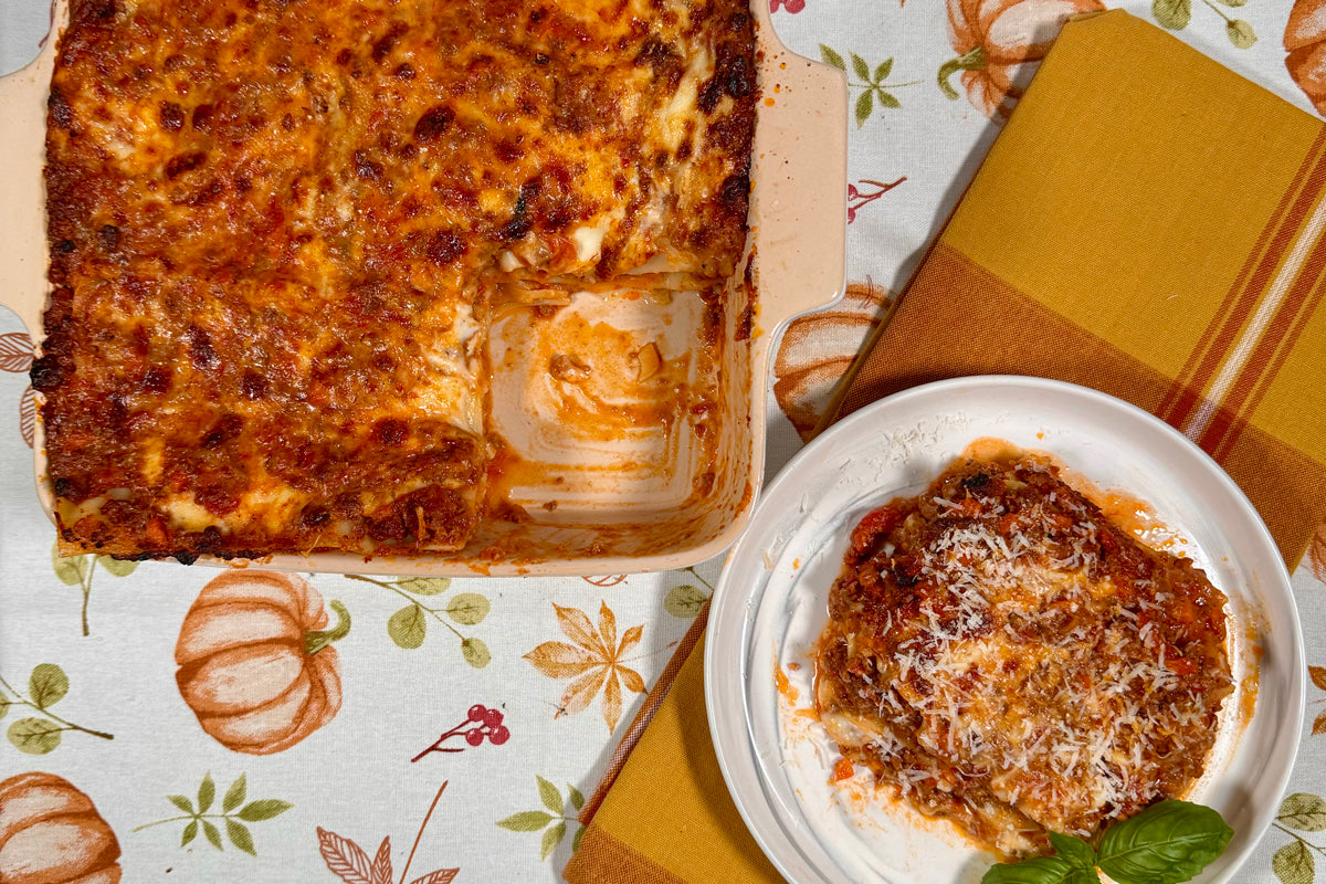 Baked lasagna with ragù and melted cheese in a ceramic baking dish, one serving on a white plate with basil on a pumpkin-print tablecloth