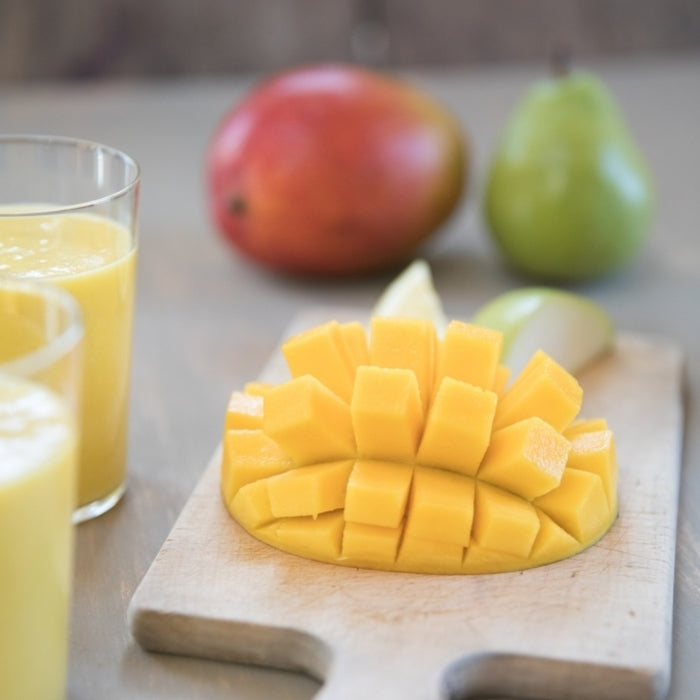 Two glasses of pear-mango smoothie beside a cubed mango on a wooden cutting board with a whole pear and mango blurred in the background