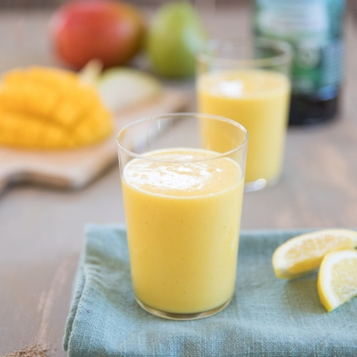 Pear-mango smoothie in a clear glass on a blue napkin, with lemon wedges and sliced mango in the blurred background