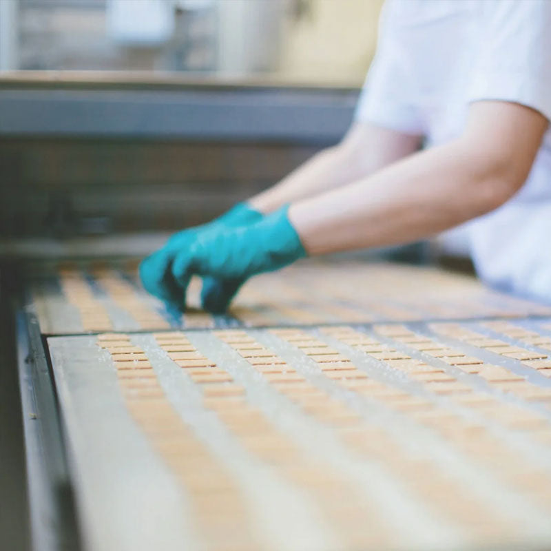 Worker wearing teal gloves arranging small rectangular crackers on a conveyor belt in a production line