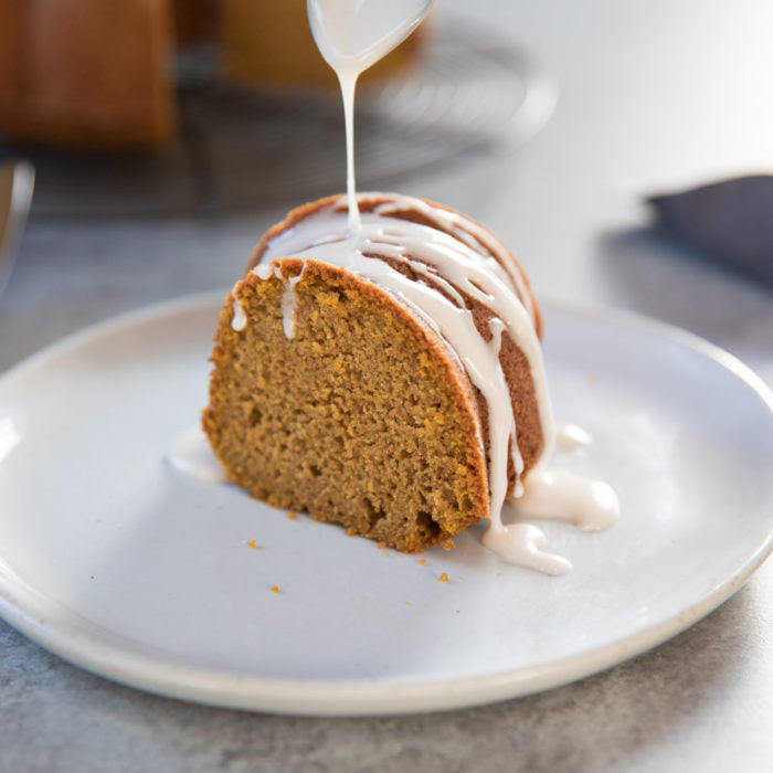 Front view of an Einkorn pumpkin spice bundt cake slice showcasing vanilla glaze being drizzled on a white plate