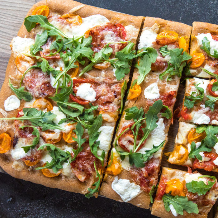 Top down view of sliced Einkorn whole wheat focaccia showcasing roasted red and yellow cherry tomatoes, dollops of ricotta, and fresh arugula on a dark baking surface
