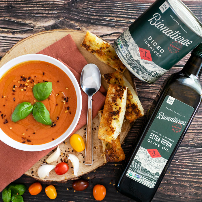 Bowl of rustic tomato soup garnished with basil, staged with grilled bread and Bionaturae tomatoes and olive oil