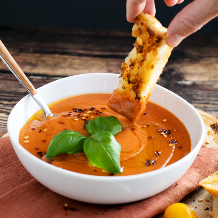 Top-down view of rustic tomato soup in a white bowl topped with basil, chili flakes and olive oil, with grilled bread