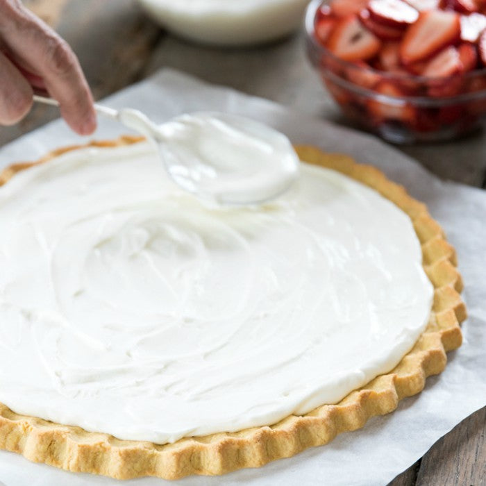 Hands spreading creamy frosting over a baked 12-inch einkorn sugar cookie crust on parchment, bowl of sliced strawberries nearby