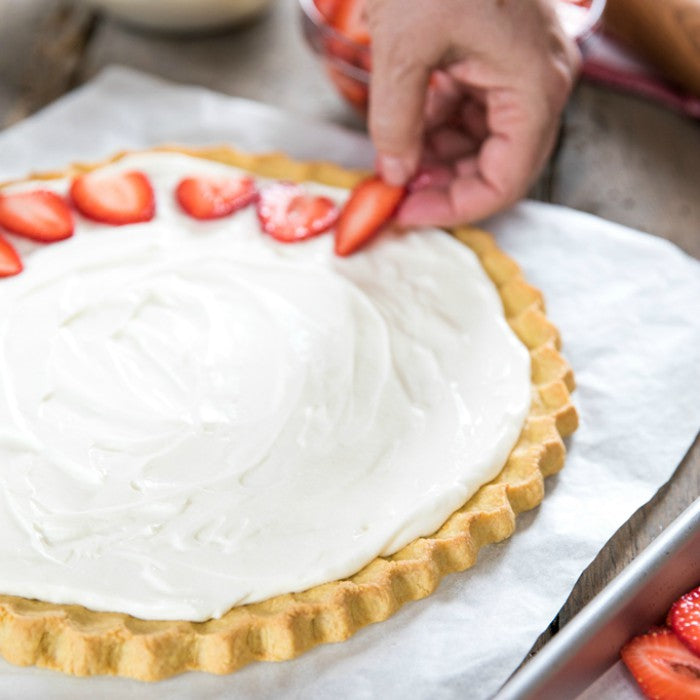 Hand arranging sliced strawberries on an Einkorn sugar cookie fruit pizza with cream cheese frosting on parchment paper