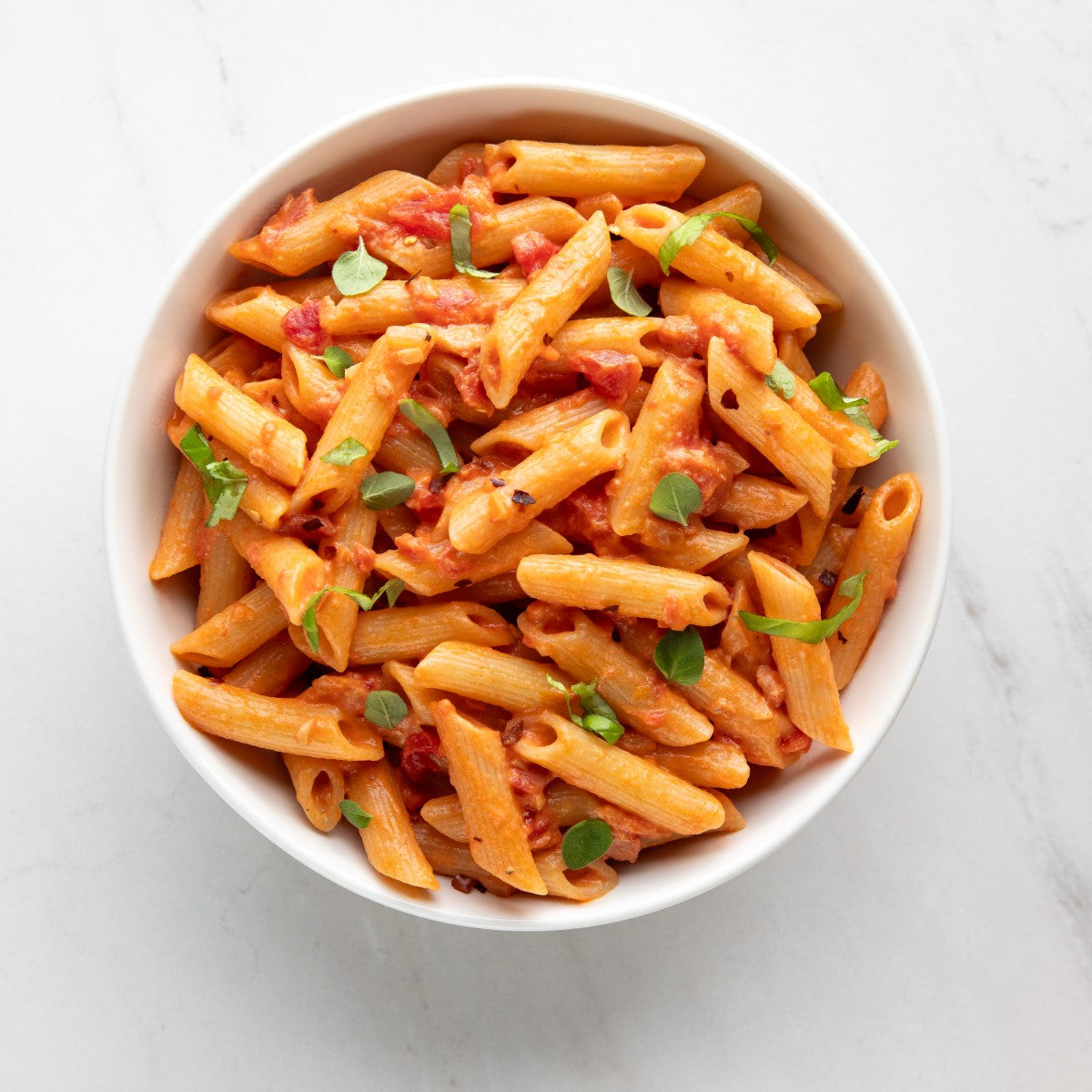 Bowl of cooked organic sourdough penne in tomato-based sauce, garnished with herbs on a white marble surface