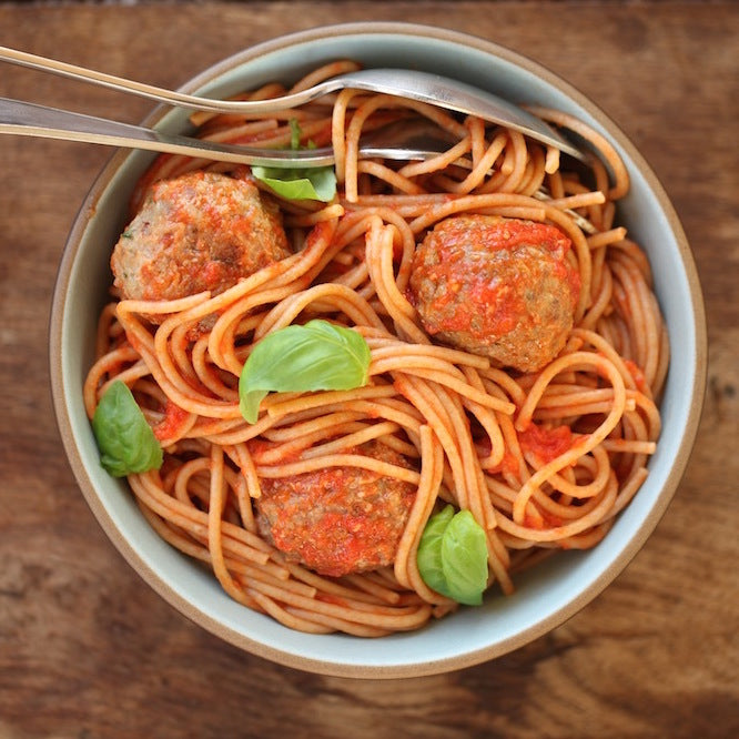 Bowl of organic einkorn whole wheat spaghetti in tomato sauce with three meatballs and basil on a wooden table