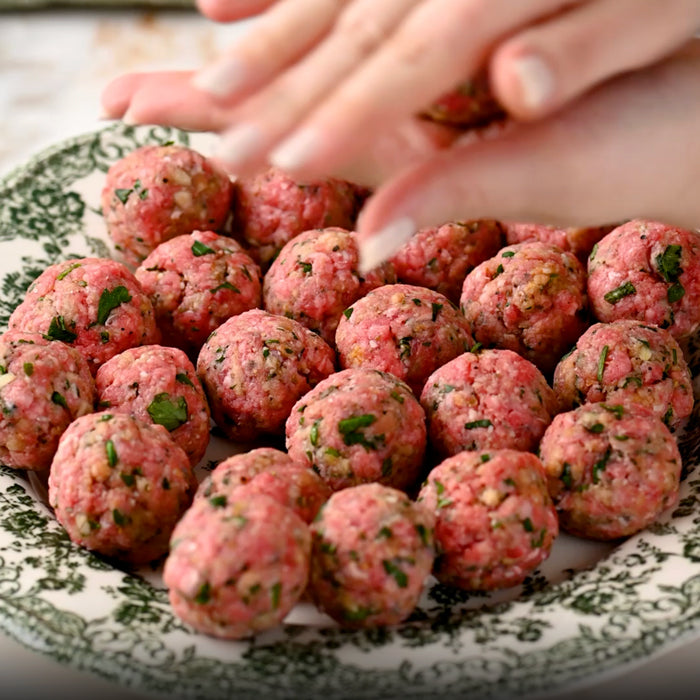 Hands shaping small seasoned Italian meatballs on a decorative plate, ready for slow cooking