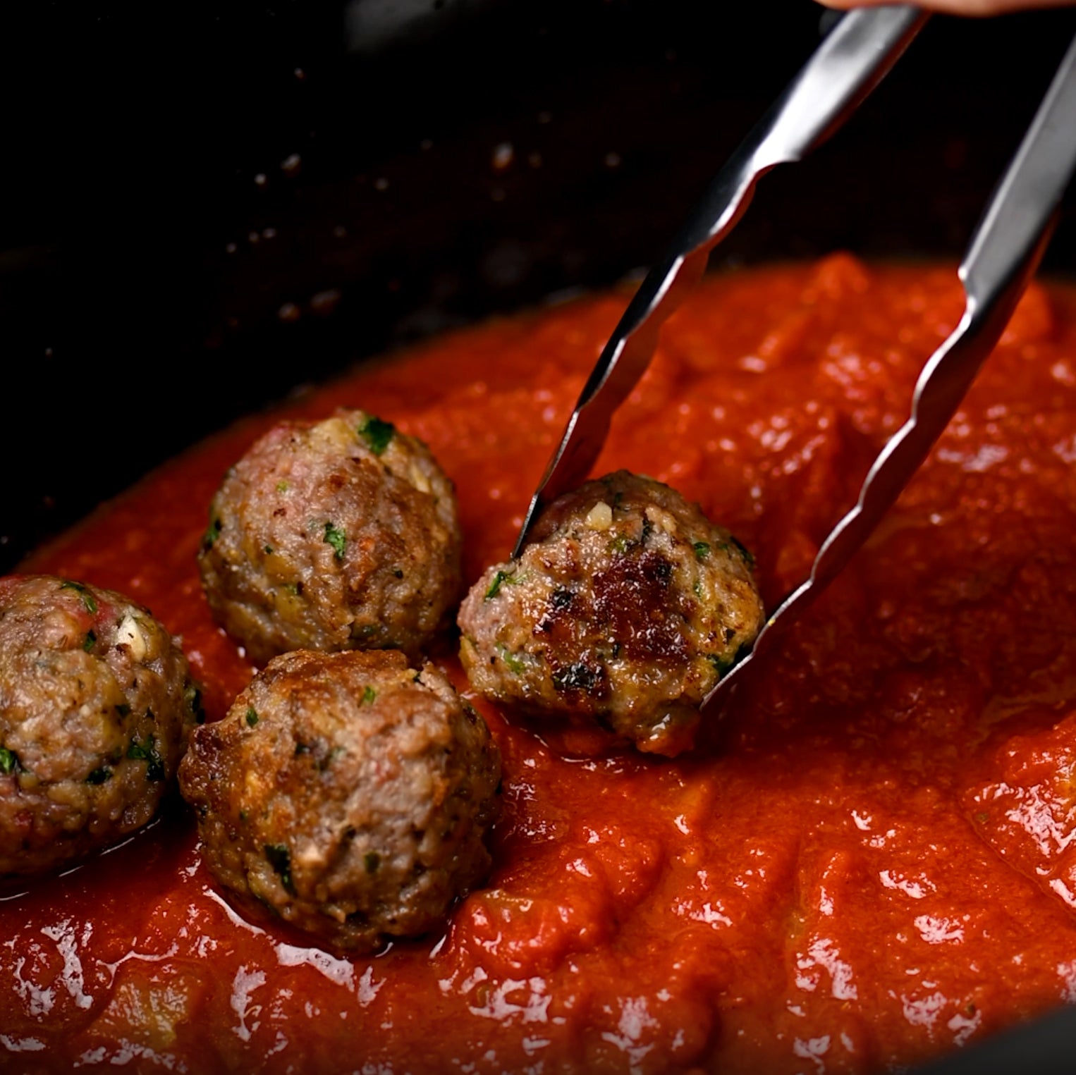 Four browned meatballs being lifted from spicy arrabbiata tomato sauce with tongs in a slow cooker