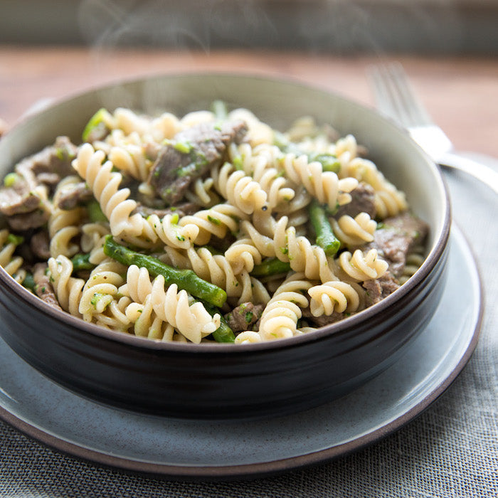Einkorn fusilli with sliced steak, asparagus, and broccoli served steaming in a ceramic bowl on a plate