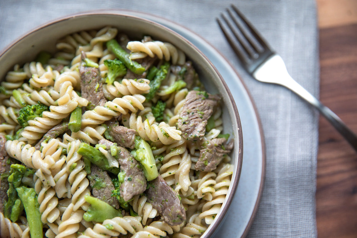 Overhead view of gluten-free fusilli with sliced steak, asparagus, and broccoli in a bowl on a cloth napkin with a fork