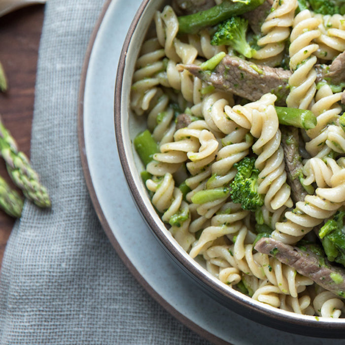 Top-down close-up of a bowl of cooked Einkorn fusilli with sliced steak, asparagus tips, and broccoli on a gray napkin