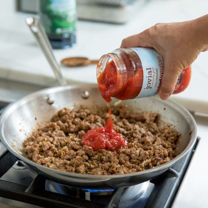 Hand pouring Jovial organic tomatoes into a skillet of browned sausage on a gas stove