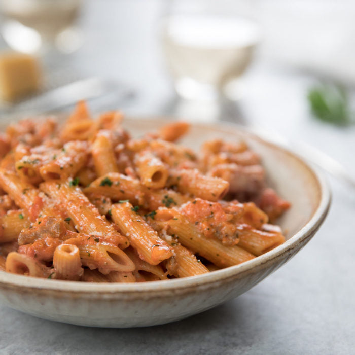 Close-up of penne pasta with Italian sausage in tomato cream sauce, garnished with parsley and grated cheese in a ceramic bowl