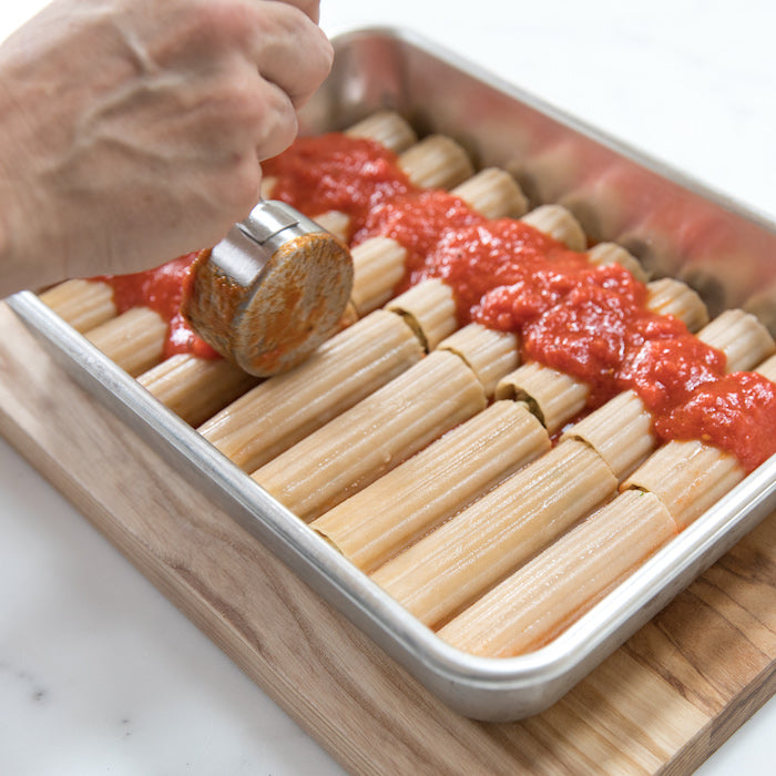 Hand spooning tomato sauce over uncooked gluten-free manicotti arranged in a baking pan on a wooden board