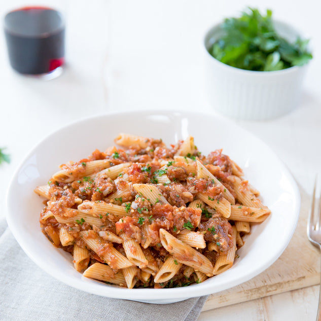 Bowl of Organic Einkorn traditional penne tossed in tomato meat sauce and garnished with parsley on a light table setting
