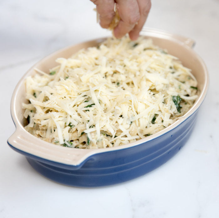 Gluten-free tuna noodle casserole in a blue oval baking dish on a marble countertop, a hand sprinkling shredded cheese on top