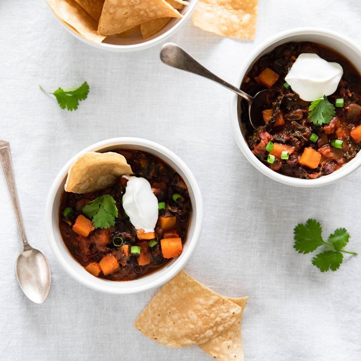 Two bowls of vegetarian borlotti bean chili topped with dollops of sour cream and cilantro, served with tortilla chips on a light tabletop